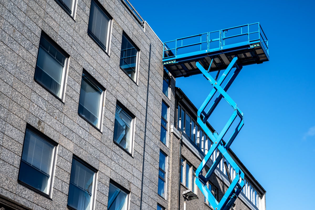 A blue electric scissor lift raised to full height beside a building